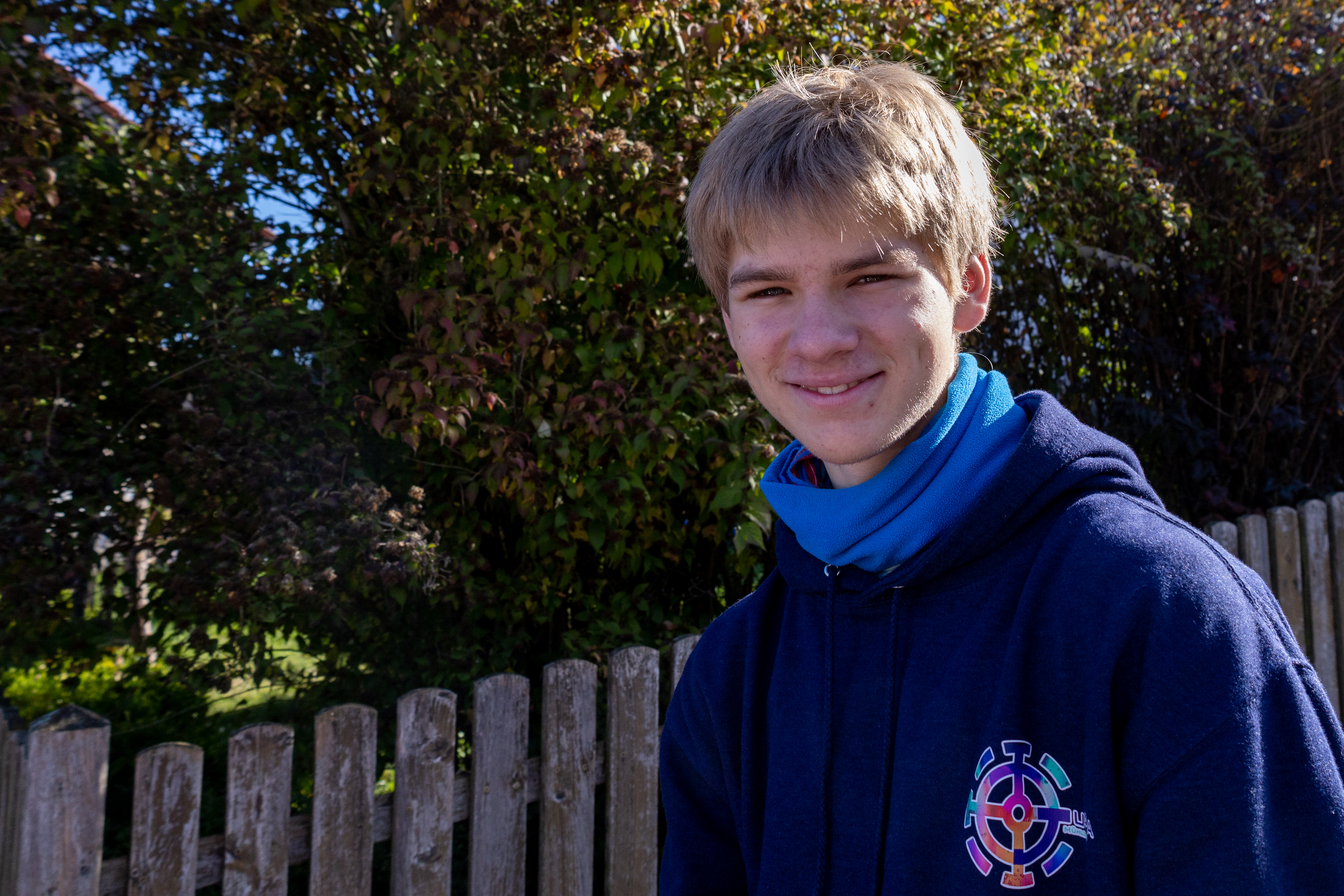 Paul Obermayer trägt einen blauen Pullover und steht vor einem Gartenzaun. 