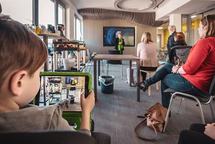 Mehrere Personen sitzen in einem Vortrag. In der hinteren Reihe sitzt ein Kind, dass den Vortrag mit seinem Tablet filmt. 