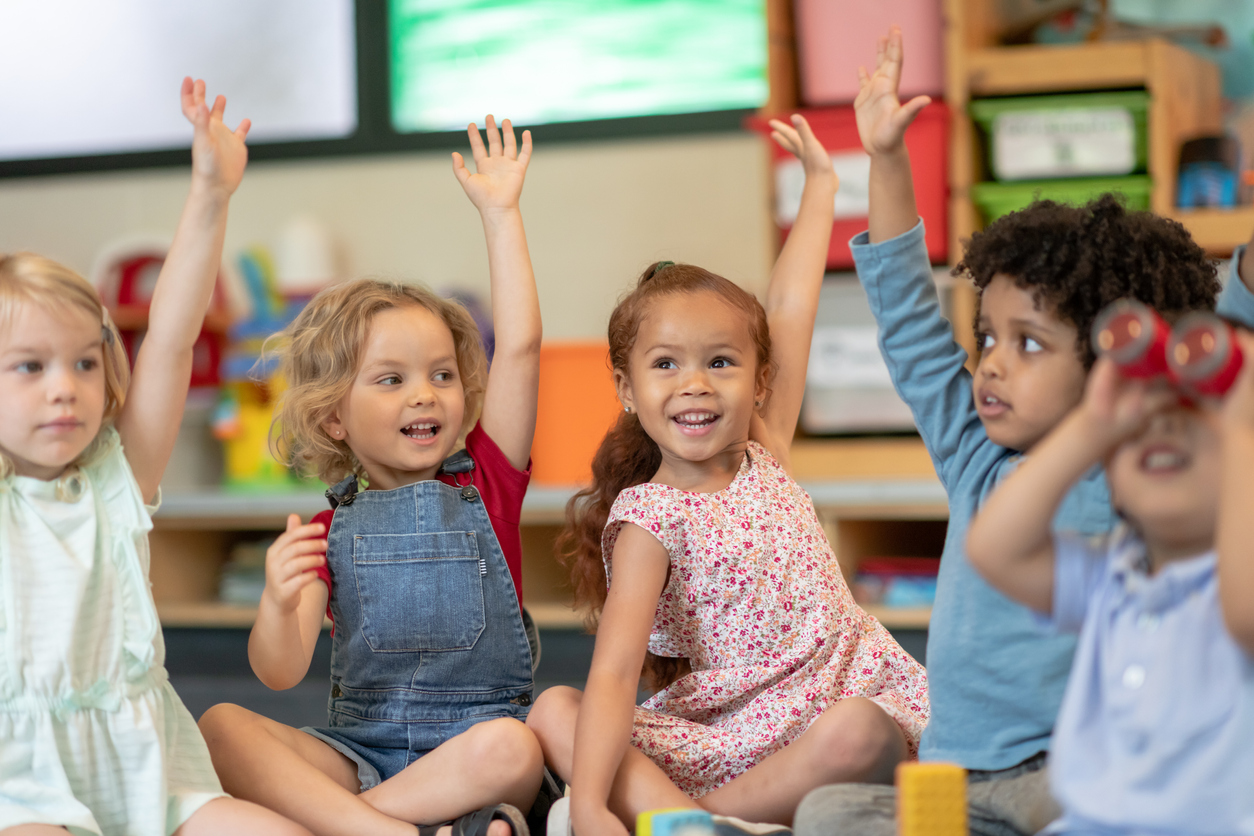 Vier Kinder sitzen nebeneinander und heben die Hände in die Höhe.